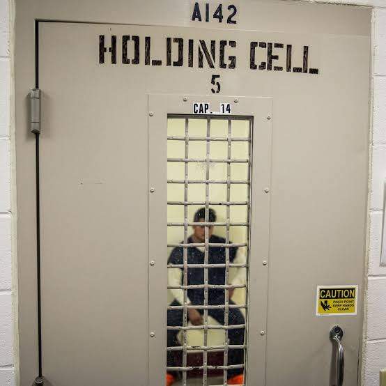 A man sits inside Holding Cell 5 at the Stewart Detention Center, behind a heavy steel door with a barred window marked CAP. 14